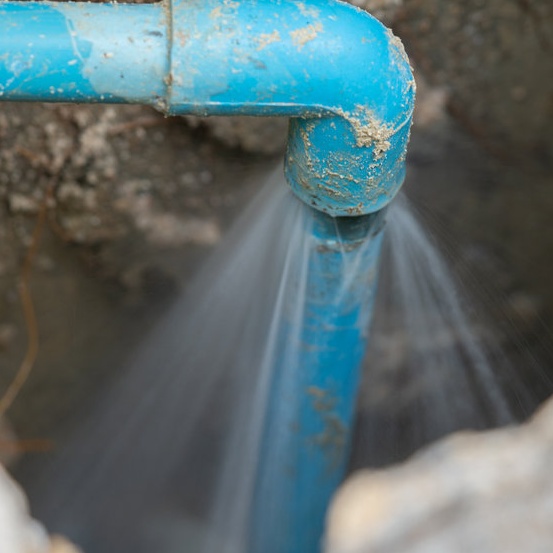 Leaking Pipes And Lines A broken blue PVC water supply pipe in an underground trench forcefully spraying a high-pressure stream of water upward from a cracked elbow joint, surrounded by wet soil and rocks.