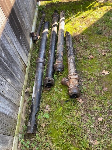 Five sections of old, corroded cast iron sewer pipe lie on grass beside a wooden fence. The pipes are heavily rusted, cracked, and covered in scale, with bell-and-spigot joints and a Y-branch visible. Moss and fallen leaves surround the debris under sunlight.