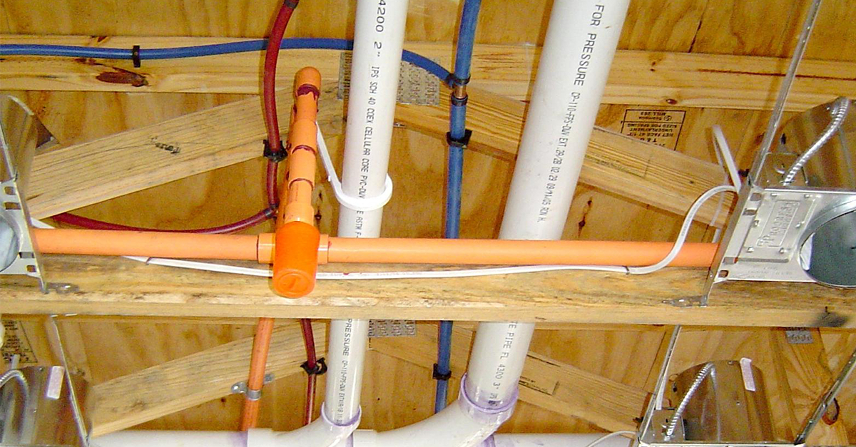 Overhead view of a plumbing rough-in between wooden floor joists: orange CPVC hot-water line with tee and drop, blue PEX cold-water line, and two white PVC vent pipes labeled "FOR PRESSURE" and "4200 2". Pipes are secured with metal clamps; HVAC duct and electrical conduit are nearby.