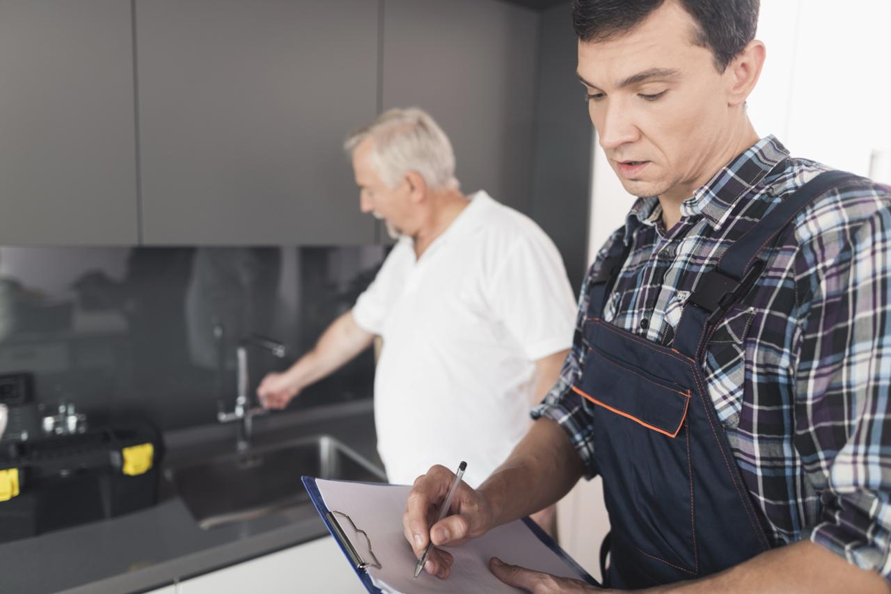 A plumber records notes on a clipboard while an older man inspects a kitchen faucet in a modern kitchen.