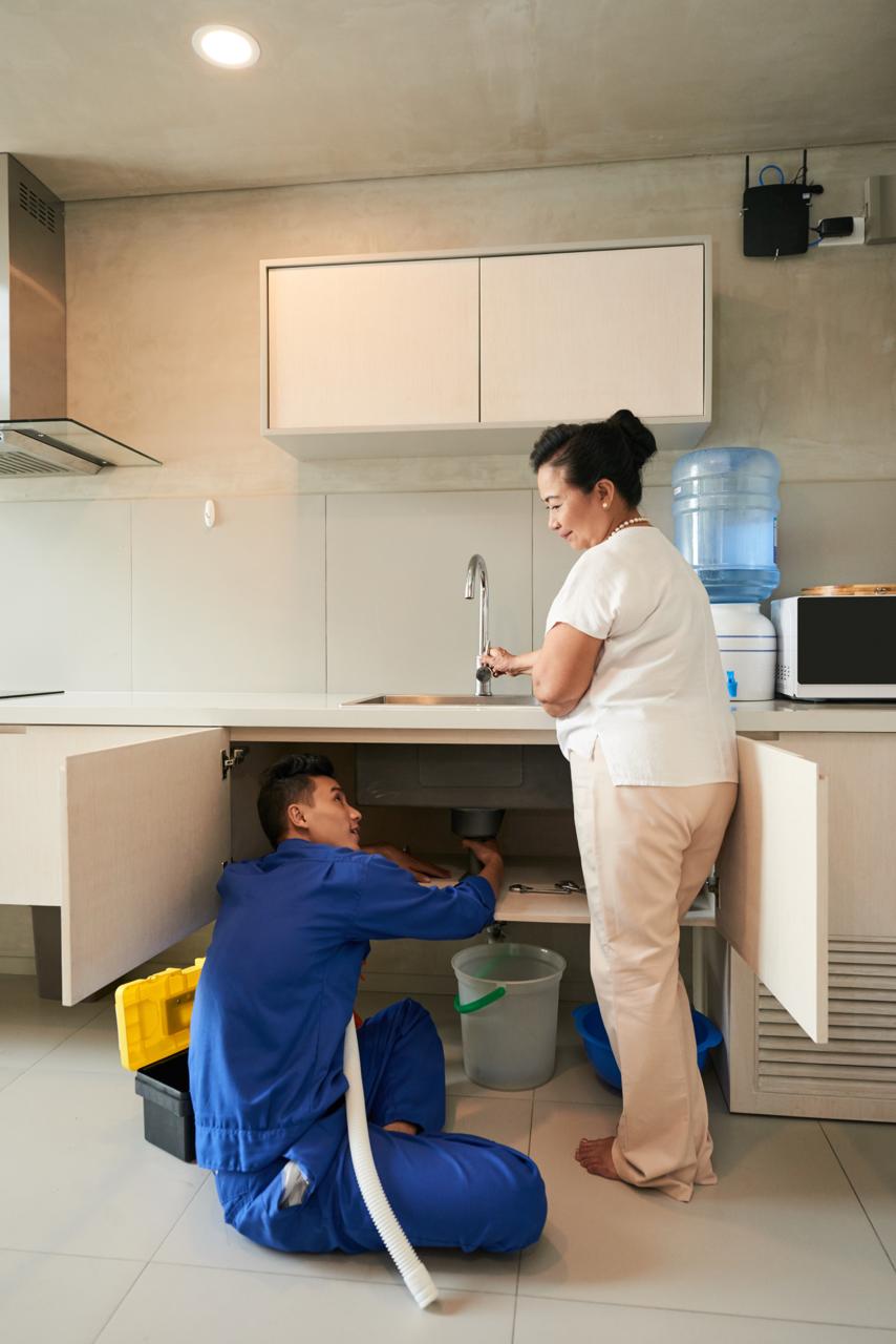 A plumber in blue coveralls fixing a kitchen sink drain while an older woman watches.